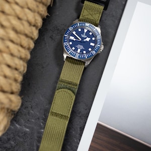 May include: A wristwatch with a blue dial and a green fabric strap. The watch features a silver bezel with white markings and a black clasp. The watch face displays the brand name "TUDOR" and the model name "PELAGOS".
