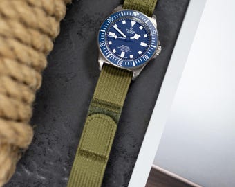 May include: A wristwatch with a blue face and a green fabric band. The watch has a silver bezel with white markings and a black clasp. The watch face displays the brand name "TUDOR" and the model name "PELAGOS".