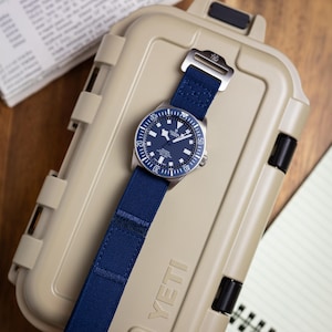 May include: A Tudor Pelagos watch with a dark blue dial and a matching blue fabric strap featuring a silver-toned metal buckle. The watch is positioned on top of a tan-coloured, rectangular, hard-shell protective case with the word YETI embossed.