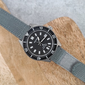 Może przedstawiać: Czarny zegarek Seiko z szarym nylonowym paskiem. Tarcza zegarka ma białe oznaczenia, a widoczny jest napis "SEIKO AUTOMATIC DIVERS 200m". Zegarek ma obracający się bezel z czarnymi oznaczeniami.