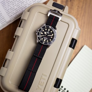 May include: A Tudor Pelagos watch with a black dial, luminous hour markers, and a rotating bezel. The watch features a black fabric strap with a thin red centre stripe and a silver-toned metal buckle, resting on a tan hard-shell storage case.