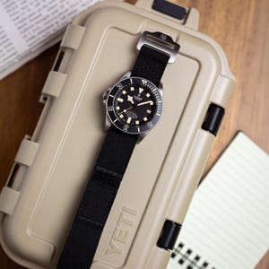 May include: A Tudor wristwatch with a black dial, luminous markers, and a rotating bezel, featuring a black fabric strap with a metal buckle, resting on a tan YETI hard-shell storage case with a secure latch mechanism and a textured surface.