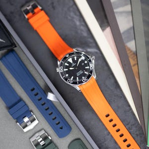 May include: A silver wristwatch with a black dial and an orange rubber strap. Additional watch straps in blue and green are visible. The watch has a black rotating bezel and white markings. The watch is displayed on a gray surface.