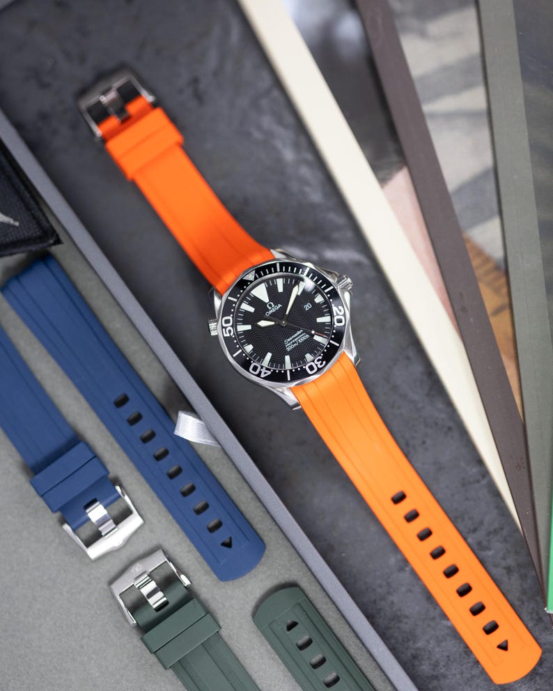 May include: A silver wristwatch with a black dial and an orange rubber strap. Additional watch straps in blue and green are visible. The watch has a black rotating bezel and white markings. The watch is displayed on a gray surface.