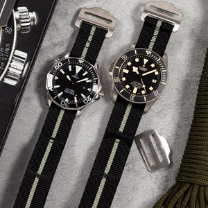May include: Two wristwatches with black and olive green striped straps. One watch features an Omega logo, the other a Tudor logo. Additional watch straps and accessories are visible on a grey surface.