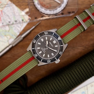 Peut inclure: Une montre Seiko noire et argentée avec un bracelet en nylon vert et rouge. La montre a un cadran noir avec des marques blanches et une fenêtre de date à 3 heures. Le bracelet a une bande rouge au centre.
