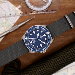 May include: A blue and silver wristwatch with a black nylon strap. The watch face has a blue dial with white markings and a silver TUDOR logo.