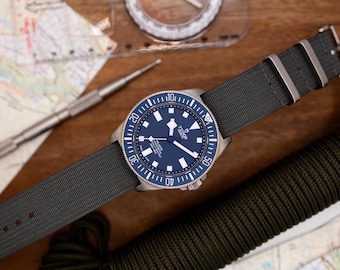 Könnte beinhalten: Eine blaue und silberne Armbanduhr mit einem schwarzen Nylonarmband. Das Zifferblatt der Uhr hat ein blaues Zifferblatt mit weißen Markierungen und ein silbernes TUDOR-Logo.