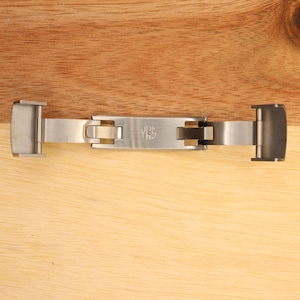 May include: A silver metal watch band clasp with a push button release. The clasp has a logo that says "JHS" on it.