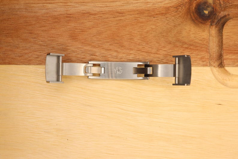 May include: A silver metal watch band clasp with a push button release. The clasp has a logo that says "JHS" on it.