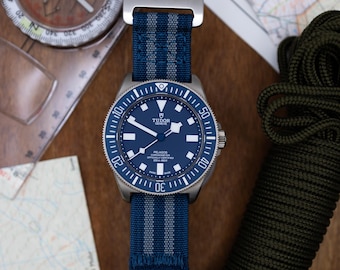 May include: A blue and silver wristwatch with a black face and white markings. The watch has a blue and gray striped nylon strap with a silver buckle. The watch face has the words "TUDOR GENEVE" and "PELAGOS" printed on it.