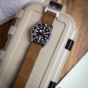May include: A Tudor Pelagos watch with a black dial, white hour markers, and a rotating bezel, featuring a brown fabric strap with a metal buckle. The watch is positioned on a tan hard-shell case with the embossed text YETI on the front surface.