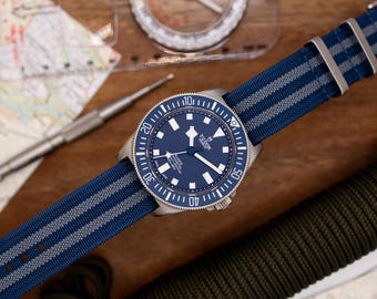 May include: A blue and silver wristwatch with a blue face and a navy blue and gray striped nylon strap. The watch has a rotating bezel with white markings and the word "TUDOR" printed on the face.