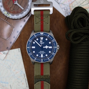 Può includere: Un orologio da polso in acciaio inossidabile con quadrante blu e cinturino in nylon a righe verdi e rosse. L'orologio è presentato con una bussola e una mappa, evocando un tema avventura.