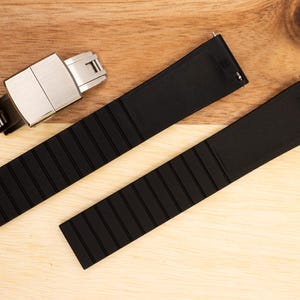 Puede incluir: Dos correas de reloj de caucho negro con una hebilla plateada. Las correas tienen una textura estriada.