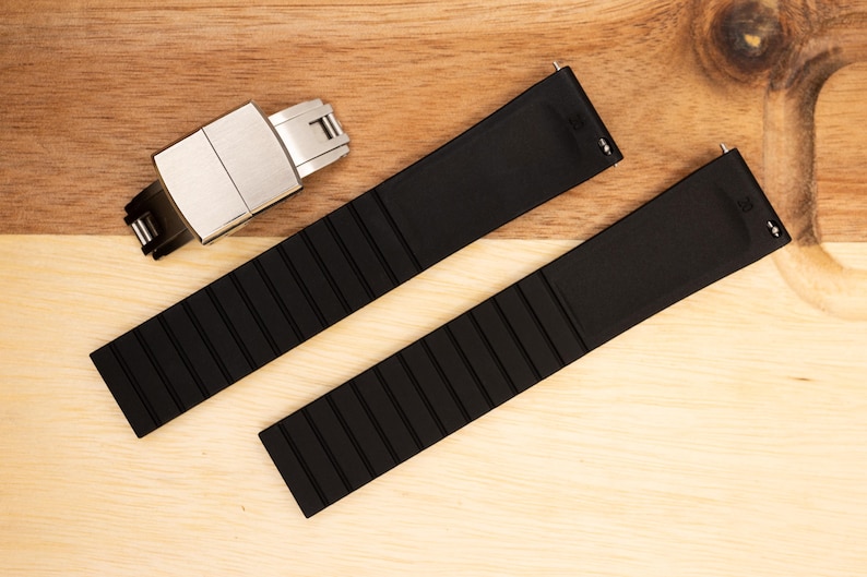 Puede incluir: Dos correas de reloj de caucho negro con una hebilla plateada. Las correas tienen una textura estriada.