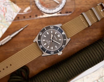 Könnte beinhalten: Eine silberne Armbanduhr mit schwarzem Zifferblatt und einem braunen Nylonarmband. Die Uhr hat ein Datumsfenster und eine drehbare Lünette.