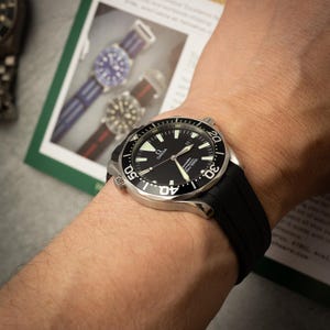 May include: A black Omega wristwatch with a black rubber strap is shown on a wrist. The watch has a black dial with white markings and a silver bezel. The watch is approximately 1.7 inches in diameter.