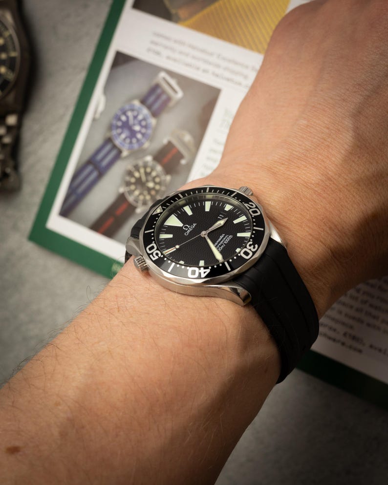 May include: A black Omega wristwatch with a black rubber strap is shown on a wrist. The watch has a black dial with white markings and a silver bezel. The watch is approximately 1.7 inches in diameter.