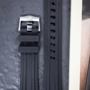 May include: A black rubber watch strap with a stainless steel buckle. The strap has a ribbed design and a series of holes for adjustment. The buckle is silver and has the brand name engraved on it.