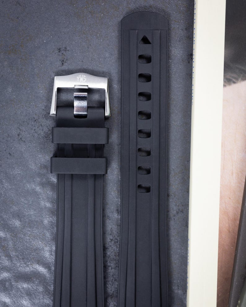 May include: A black rubber watch strap with a stainless steel buckle. The strap has a ribbed design and a series of holes for adjustment. The buckle is silver and has the brand name engraved on it.