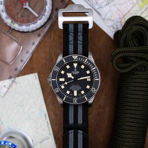 May include: A black and silver wristwatch with a black nylon strap and a silver buckle. The watch face has a black background with white markings and a silver TUDOR GENEVE logo. The watch is surrounded by a black bezel with white markings.