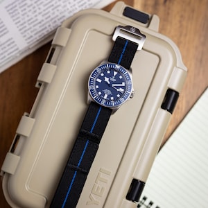 May include: A Tudor Pelagos watch with a blue dial and a black fabric strap featuring a thin blue centre stripe. The watch has a rotating bezel with white markers and is resting on a tan, rectangular, hard-shell protective case with YETI branding.