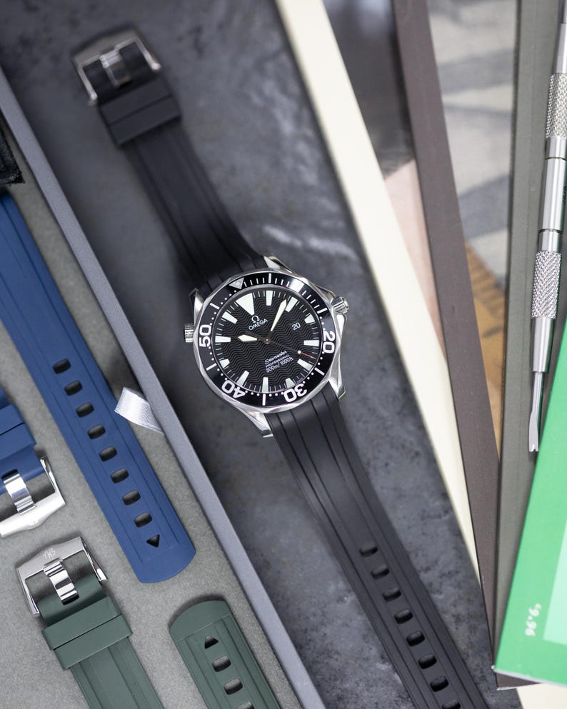 May include: A black Omega Seamaster watch with a black rubber strap. The watch has a black dial with white markings and a silver bezel. Additional blue and green watch straps are visible.