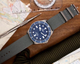 Könnte beinhalten: Eine Armbanduhr mit blauem Zifferblatt, silberner Lünette und grauem Nylonarmband. Das Zifferblatt der Uhr hat silberne Zeiger und Markierungen, und das Wort "TUDOR" ist auf dem Zifferblatt aufgedruckt.