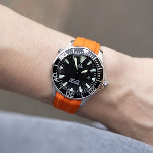 May include: A stainless steel wristwatch with a black dial and orange band. The watch face features white markers and hands, with the brand name "OMEGA" visible. The orange band is made of a flexible material. The watch is designed for water resistance.
