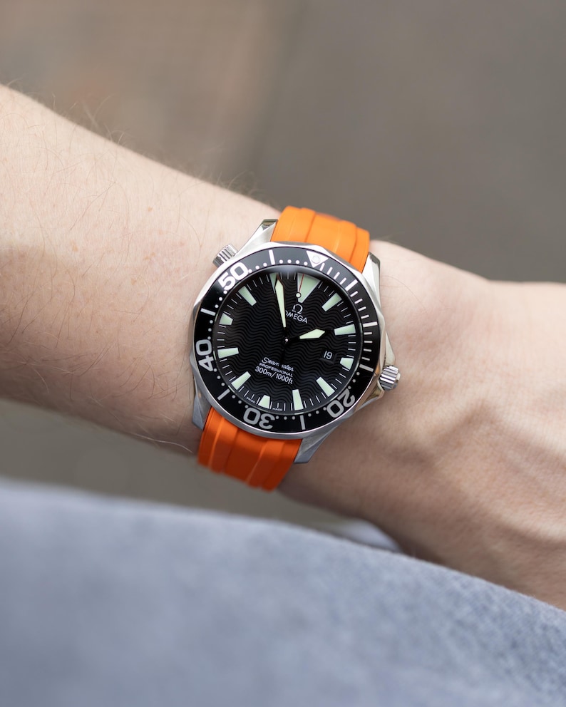 May include: A stainless steel wristwatch with a black dial and orange band. The watch face features white markers and hands, with the brand name "OMEGA" visible. The orange band is made of a flexible material. The watch is designed for water resistance.