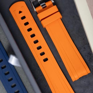 May include: Bright orange watch strap with a silver buckle. The strap features a series of rectangular holes and a textured surface. The watch band is made of a flexible material.