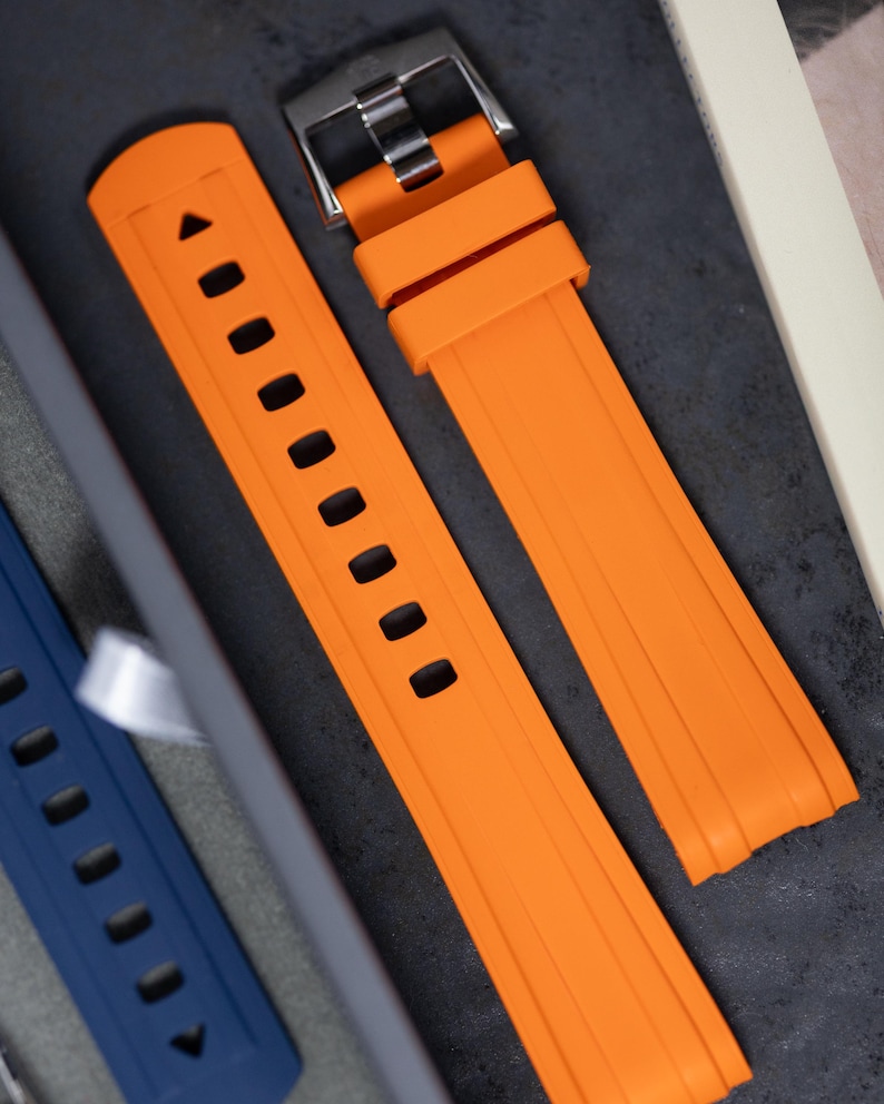 May include: Bright orange watch strap with a silver buckle. The strap features a series of rectangular holes and a textured surface. The watch band is made of a flexible material.