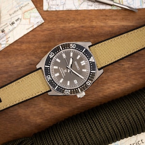 May include: A silver watch with a black face and a beige canvas strap. The watch has a rotating bezel with numbers from 10 to 50. The watch face has the word "SEIKO" and the text "Automatic 23 Jewels"