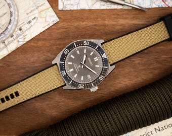 May include: A silver watch with a black face and a beige canvas strap. The watch has a rotating bezel with numbers from 10 to 50. The watch face has the word "SEIKO" and the text "Automatic 23 Jewels"