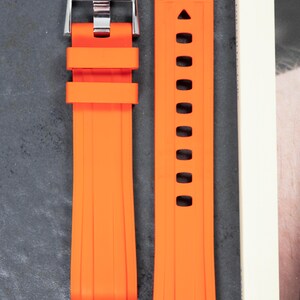 May include: Bright orange watch strap with a silver buckle. The strap is made of a flexible material and features a series of holes for adjustment. The buckle is engraved with the letters "YWS".
