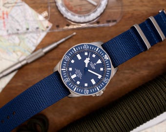 May include: A blue and silver wristwatch with a blue nylon strap. The watch face has a white dial with silver hands and markers. The watch has a rotating bezel with numbers and markings.