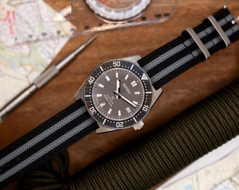 May include: A black and gray striped nylon watch strap with a silver watch face. The watch face has black markings and a date window at the 3 o'clock position. The watch is a Seiko Automatic Diver's watch.
