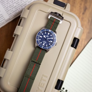 May include: A Tudor Pelagos watch with a blue dial and a rotating bezel, featuring a green fabric strap with a thin red vertical stripe running down the centre. The watch is positioned on top of a tan, rectangular, hard-shell protective storage case.