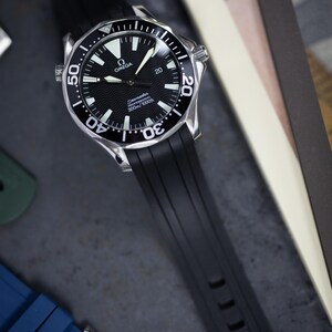 May include: A black and silver wristwatch with a black rubber strap. The watch face is black with white markers and hands, and the bezel has white markings. The watch is branded with the Omega logo and the text "Seamaster 300m/1000ft".