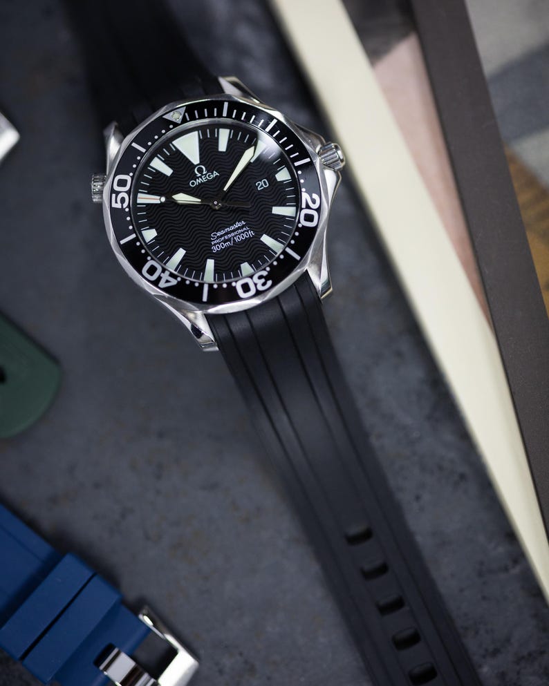 May include: A black and silver wristwatch with a black rubber strap. The watch face is black with white markers and hands, and the bezel has white markings. The watch is branded with the Omega logo and the text "Seamaster 300m/1000ft".