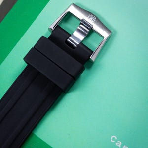 May include: Close-up of a black watch strap with a silver buckle. The buckle has the letters "JWS" engraved on it. The watch strap is made of a ribbed, textured material. The background is a light green color.