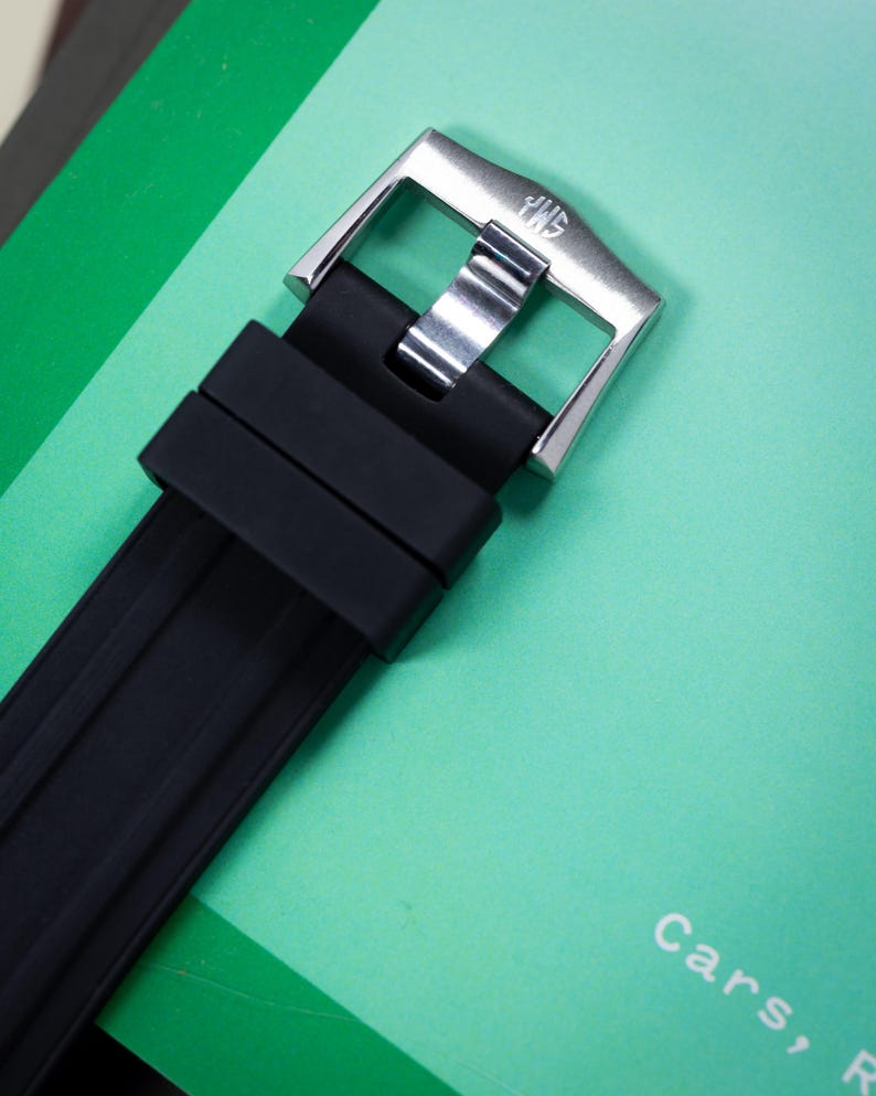 May include: Close-up of a black watch strap with a silver buckle. The buckle has the letters "JWS" engraved on it. The watch strap is made of a ribbed, textured material. The background is a light green color.