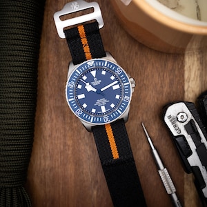 May include: A black and orange NATO watch strap with a silver buckle. The strap is on a silver wristwatch with a blue face and silver hands. The watch face has the text "TUDOR GENEVE" and "PELAGOS" printed on it.