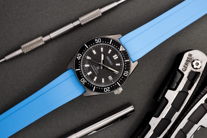 May include: A black and silver Seiko watch with a blue rubber strap. The watch has a black dial with white markings and a date window at the 3 o'clock position. The watch is on a gray surface.