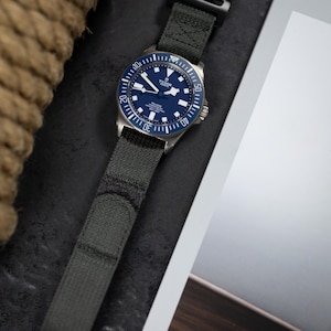 May include: A stainless steel wristwatch with a dark blue face and a green fabric strap. The watch has a rotating bezel with white markings and a black clasp. The watch face displays the brand name and other details. The strap is approximately 2.5 cm wide.