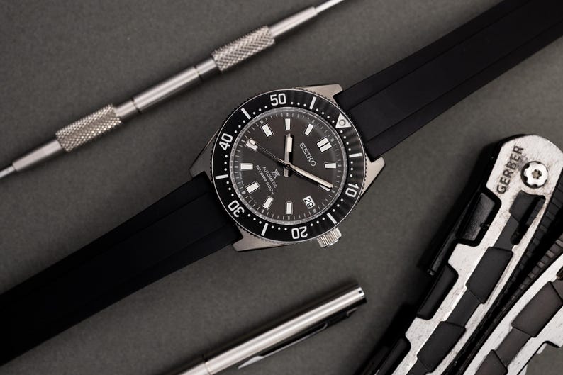 Puede incluir: Un reloj Seiko negro y plateado con una correa de caucho negro. El reloj tiene una esfera negra con marcas blancas y una ventana de fecha en la posici&oacute;n de las 3 en punto. El reloj est&aacute; rodeado por un bisel negro con marcas blancas.