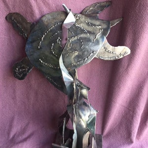 May include: A metal sculpture of a sea turtle with a textured shell and flippers. The sculpture is made of welded metal and stands on a square base. The artwork has a modern, abstract design.