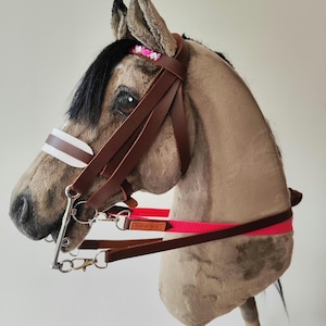 Op de afbeelding: Een pluchen stokpaard met een bruin lichaam, zwarte manen en een bruin leren hoofdstel. Het hoofdstel heeft een wit en bruin frontriem en een felroze band. Het paard heeft een houten stok als handvat en is versierd met kleine roze bloemen.