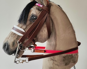 May include: A plush hobby horse with a tan body, black mane, and brown leather bridle. The bridle features a white and brown browband and a bright pink strap. The horse has a wooden stick for a handle and is decorated with small pink flowers.